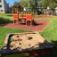 Sandbox with toy trucks and playground equipment in the background on a sunny day.