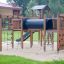 Romping Cottage wooden playground equipment with climbing nets, ladders, and slide in a park.