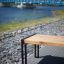 Modern wooden Thor long bench with metal legs alongside a riverside path with a blue bridge in the b
