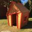 Wooden playhouse for children - Crooked House, brown with red trim, outdoor setting.