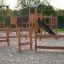 Trim trail wooden playground equipment with climbing frames and slide on sandy ground.