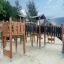 Modern wooden playground structure with slide and climbing elements, surrounded by sand and trees.