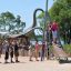 Children playing on the Silver Cableway playground equipment with a dinosaur statue in the backgroun