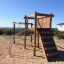 Kampinos wooden playhouse in a sandy outdoor area with climbing elements and a slide.