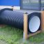 Children's play tunnel made of black corrugated tube and wooden frames on grassy area.