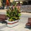 Planter Geneva IV with lush green foliage and orange flowers on a paved public square between wooden