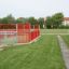 Red play area surrounded by a metal fence on green grass in a park next to a building.
