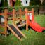 Mini play wooden playground set with a red slide and climbing ramp on a green lawn.