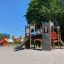 Camelot castle with slide playground structure in a park with trees and buildings in the background.