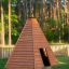 Wooden teepee-style playhouse on grass with forest background.