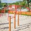 Playground equipment Somersault Double on a sandy area with red metal arches and wooden posts.